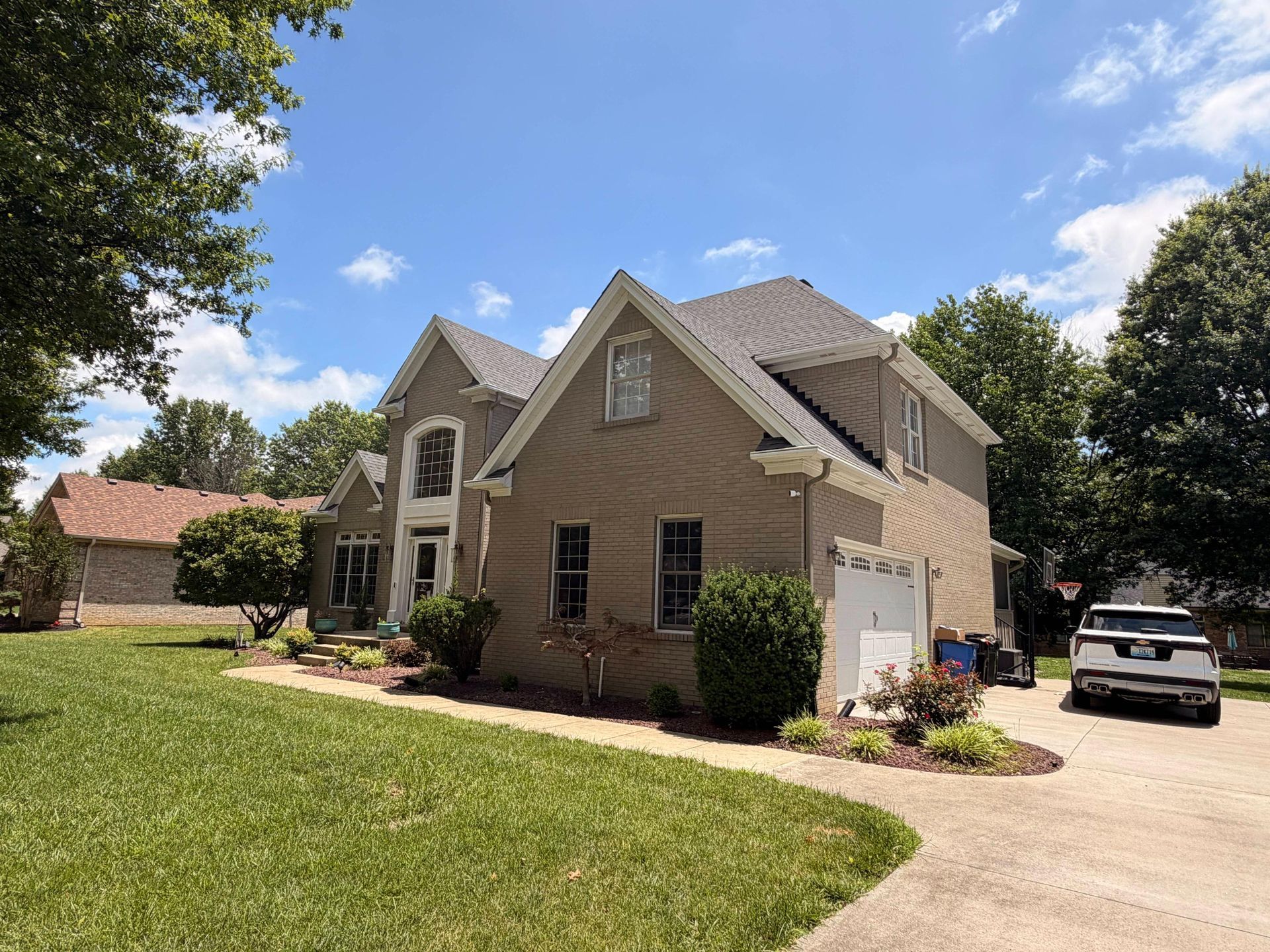 Suburban house with tan siding, front lawn, driveway, and parked car on a sunny day