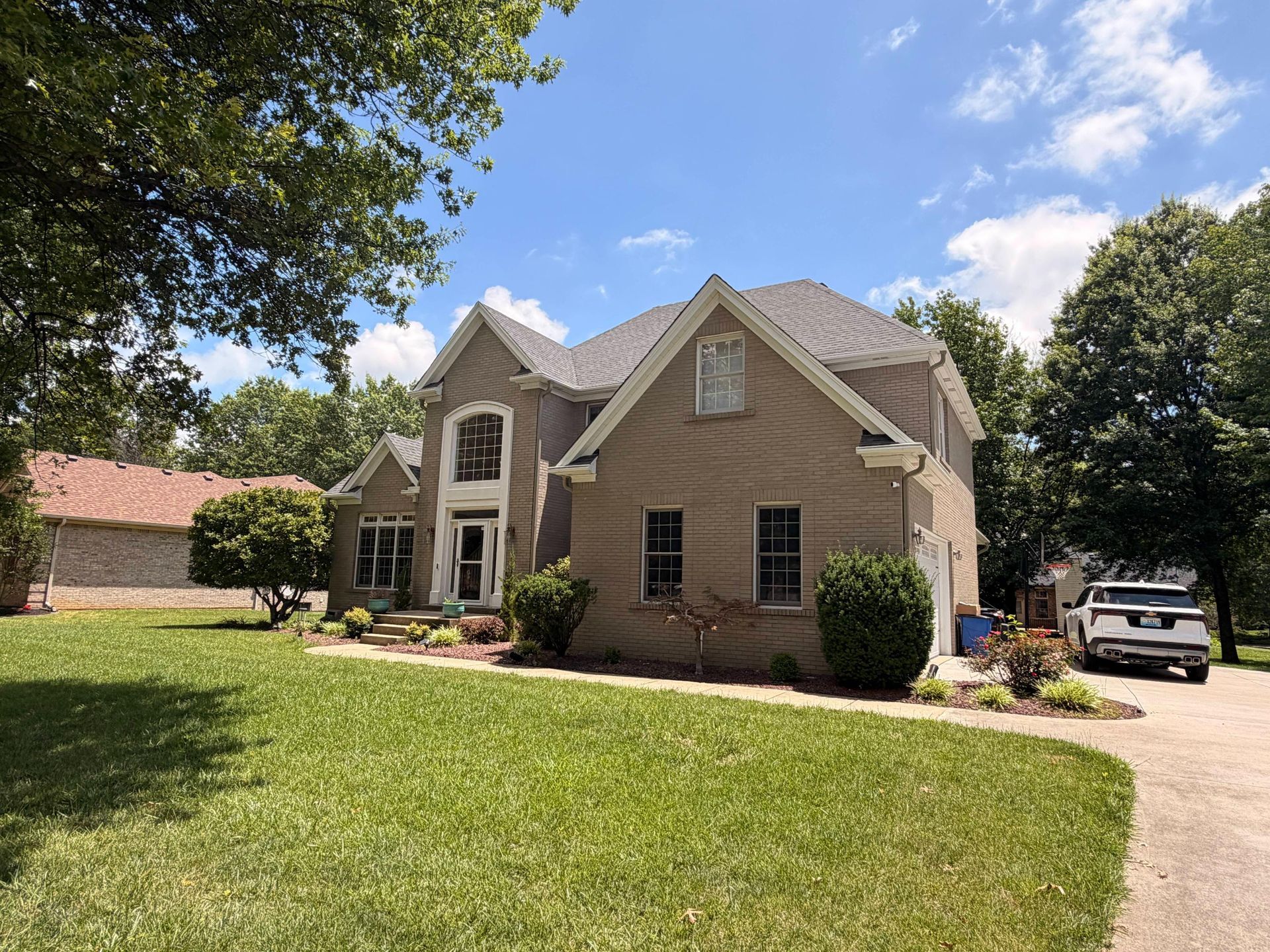 Suburban beige house with gabled roof, front lawn, and a parked SUV under a blue sky