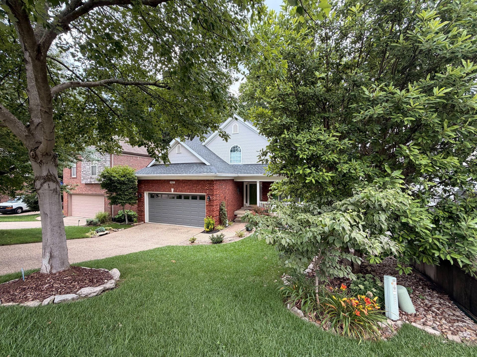 Suburban brick house with gray roof, front lawn, large tree, and flowerbed along the sidewalk