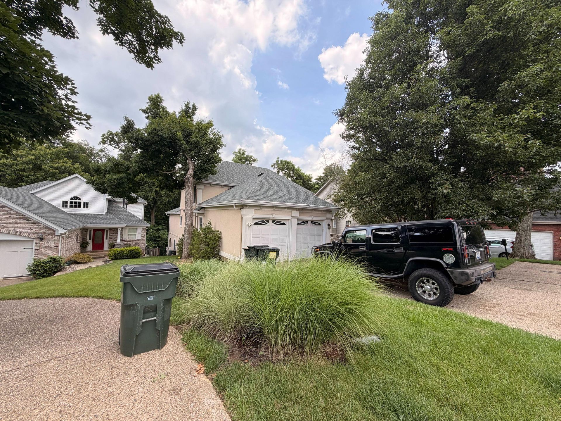 Suburban house with gravel driveway, black SUV, green trash bin, and tall trees under a partly cloudy sky