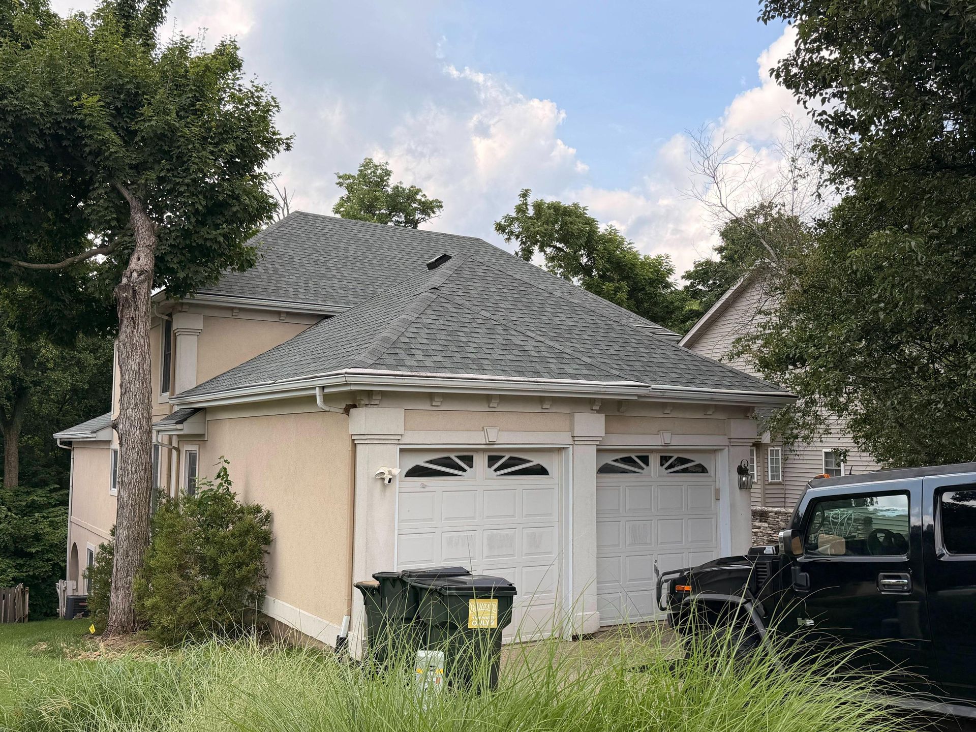Beige house with white garage doors, gray roof, and black truck parked in the driveway amid trees.