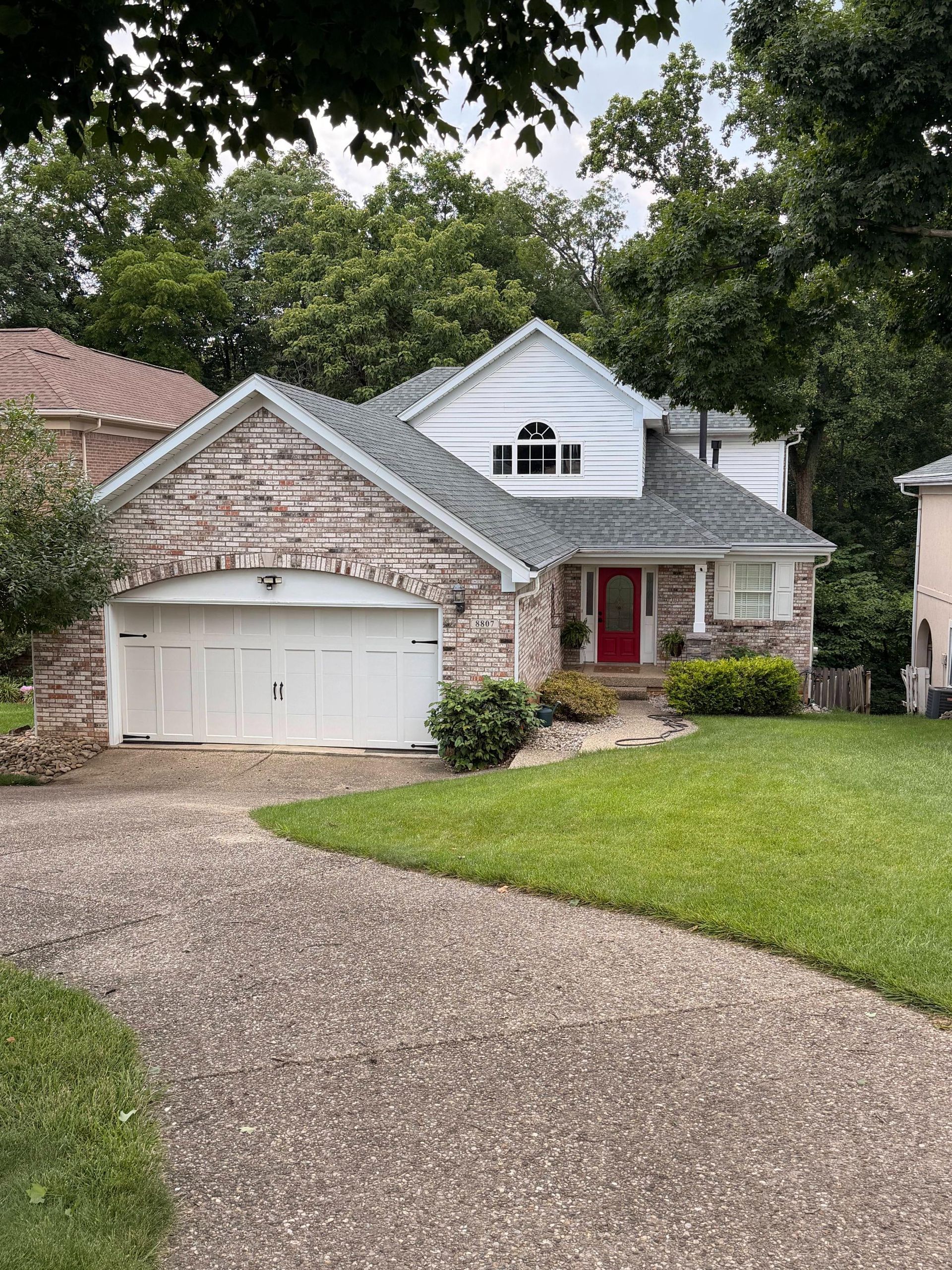 Suburban house with stone garage, gray roof, red door, and curved gravel driveway under trees