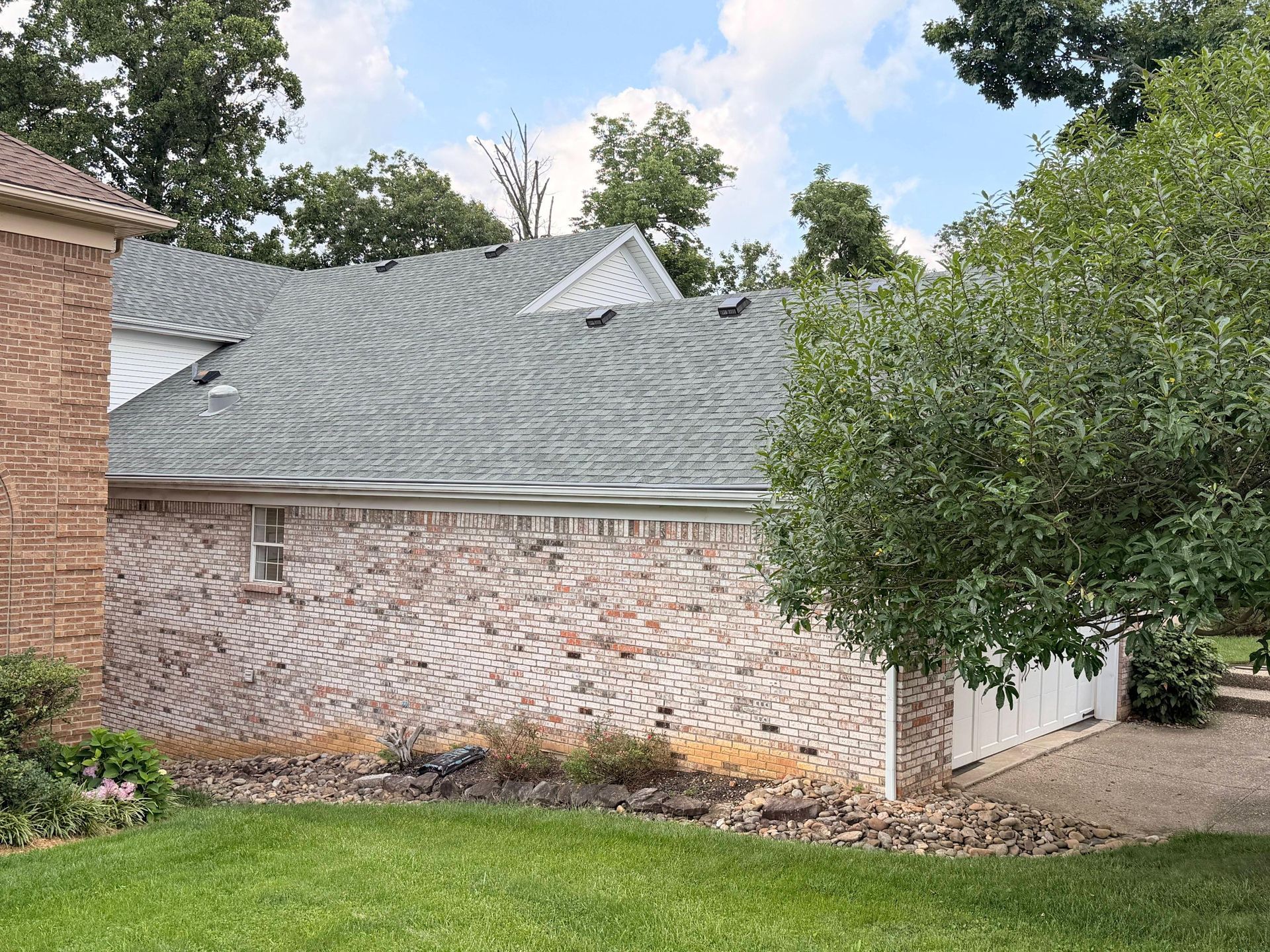Brick house with gray shingle roof, white side addition, and a leafy tree in front