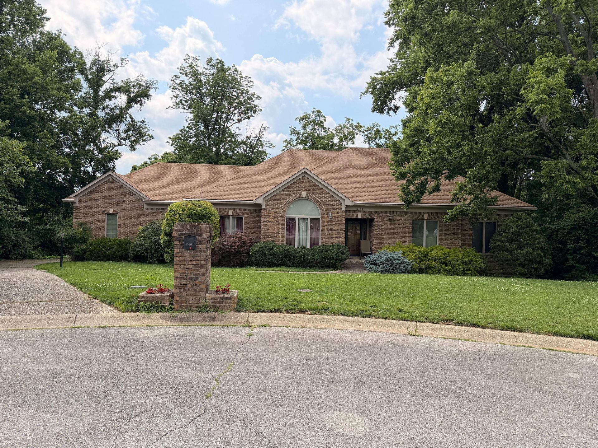Brick single-story house with a manicured lawn, trees, and a driveway under a partly cloudy sky
