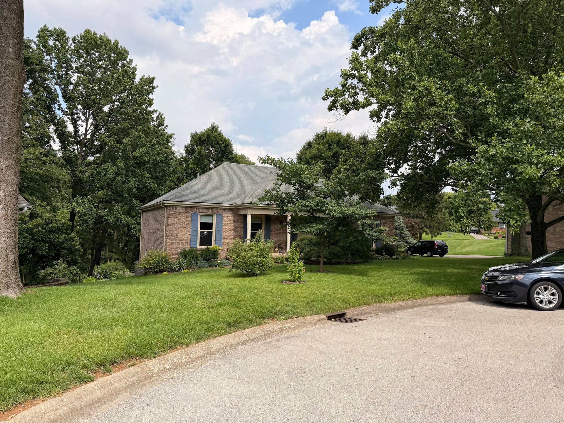 Small brick house with a green roof on a grassy corner lot, trees around it, and parked cars nearby.