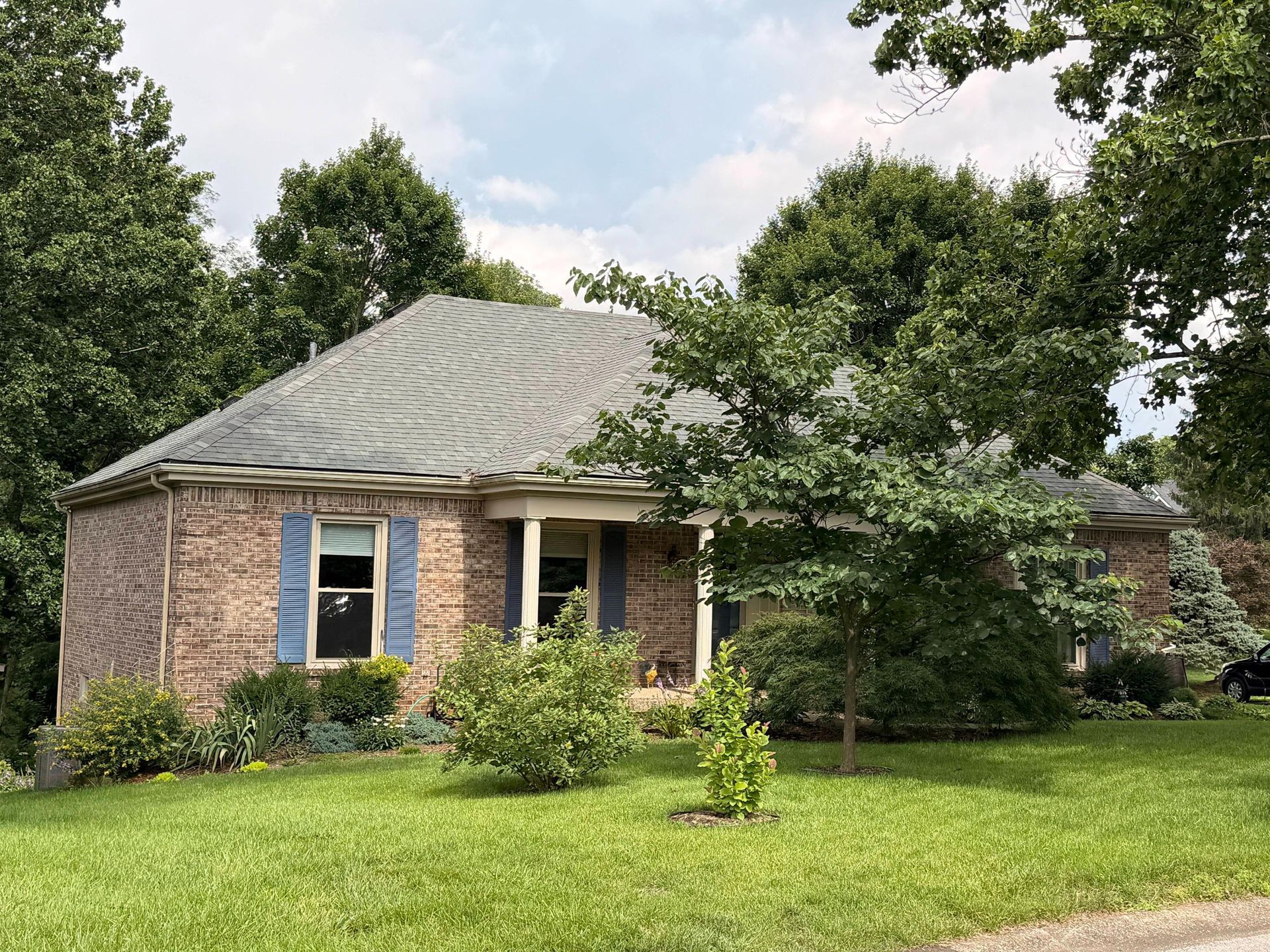 Single-story brick house with gray roof, blue shutters, and a green lawn surrounded by trees.