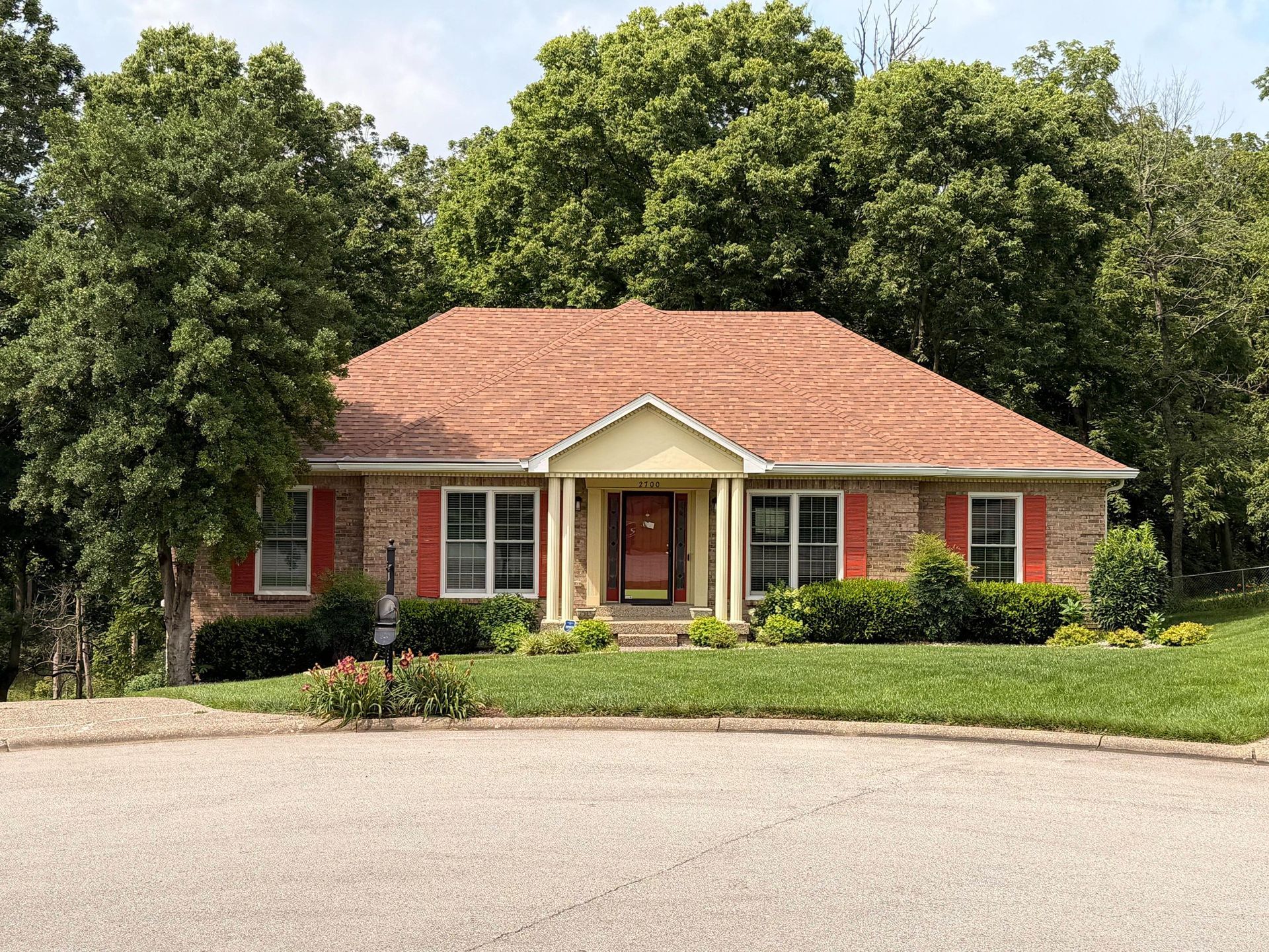 Single-story brick house with red shutters, white trim, and a red-tile roof surrounded by trees and lawn