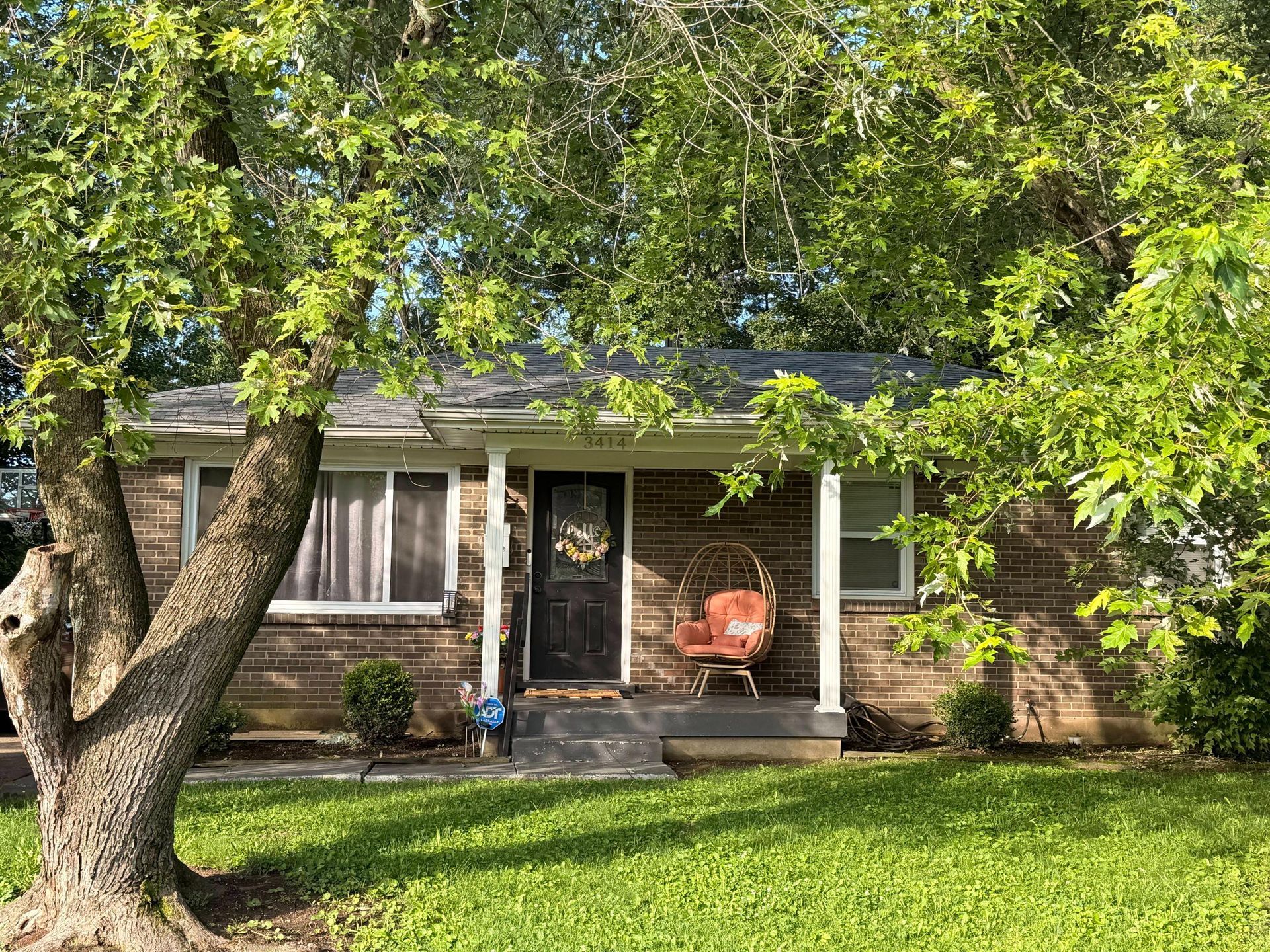 Brick house with a front porch shaded by large trees and a green lawn in the foreground