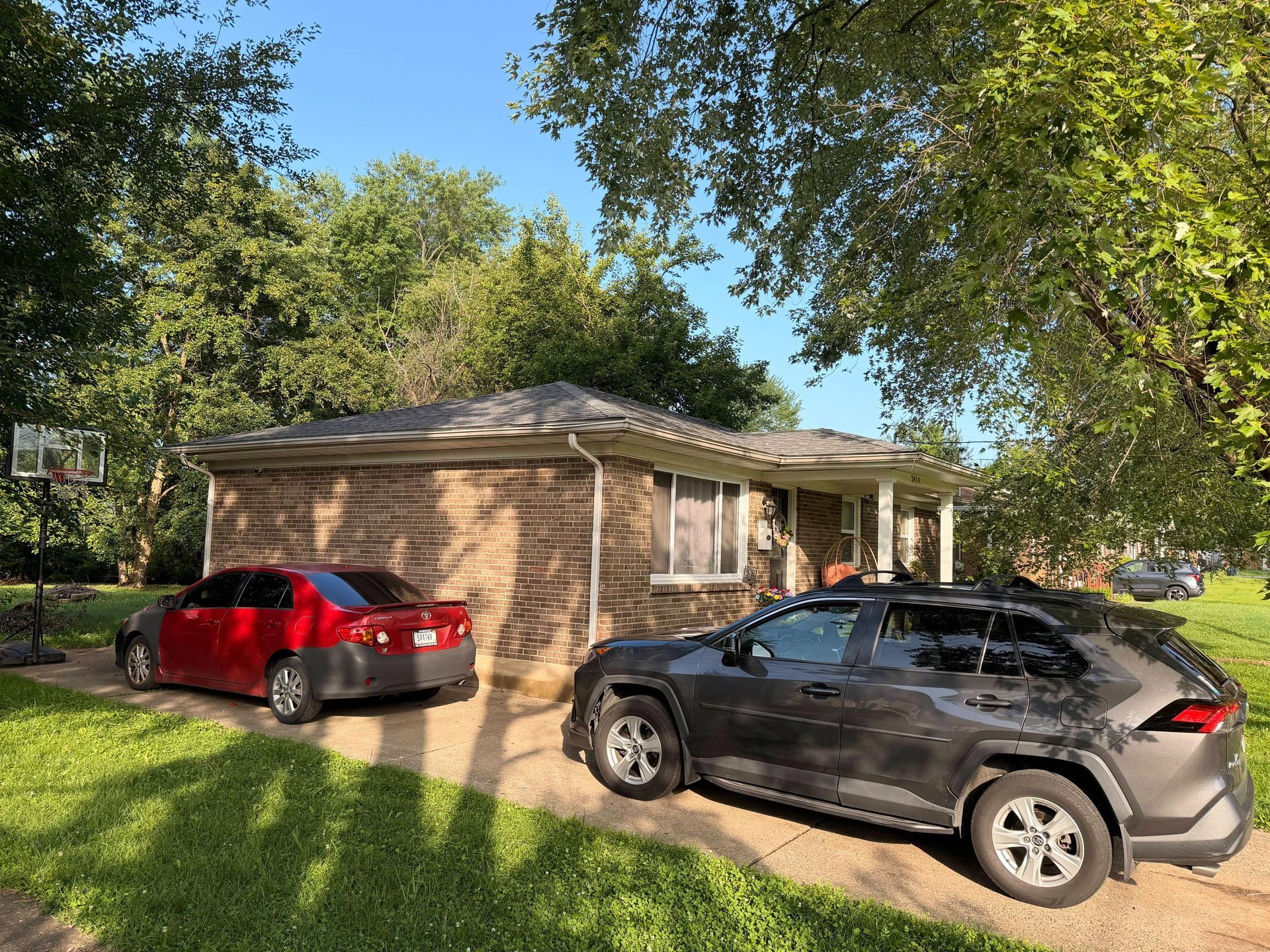 Two cars parked beside a brick house under trees on a sunny day
