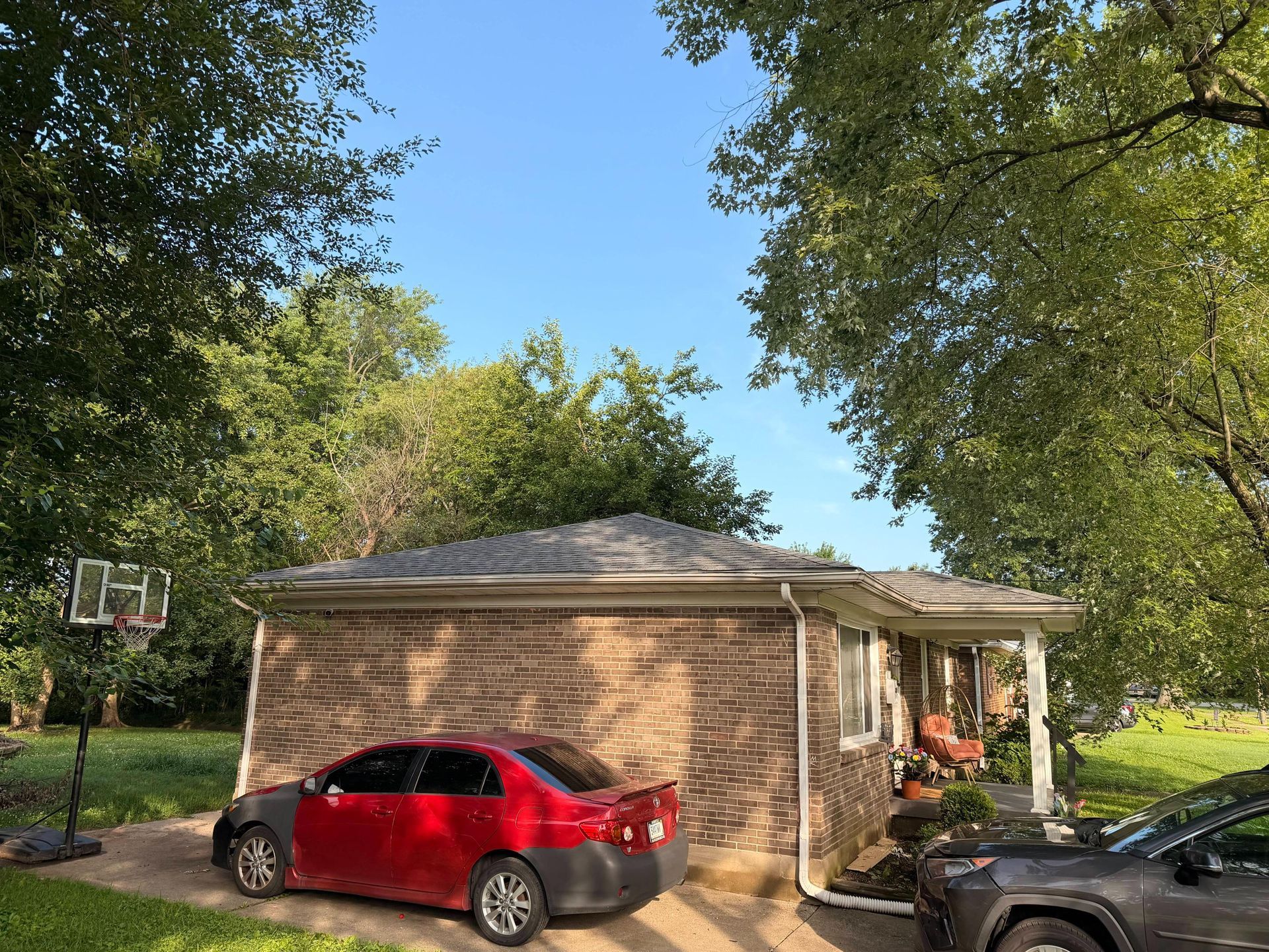 Brick house with a red car parked in front, trees overhead, and a covered porch on the right.