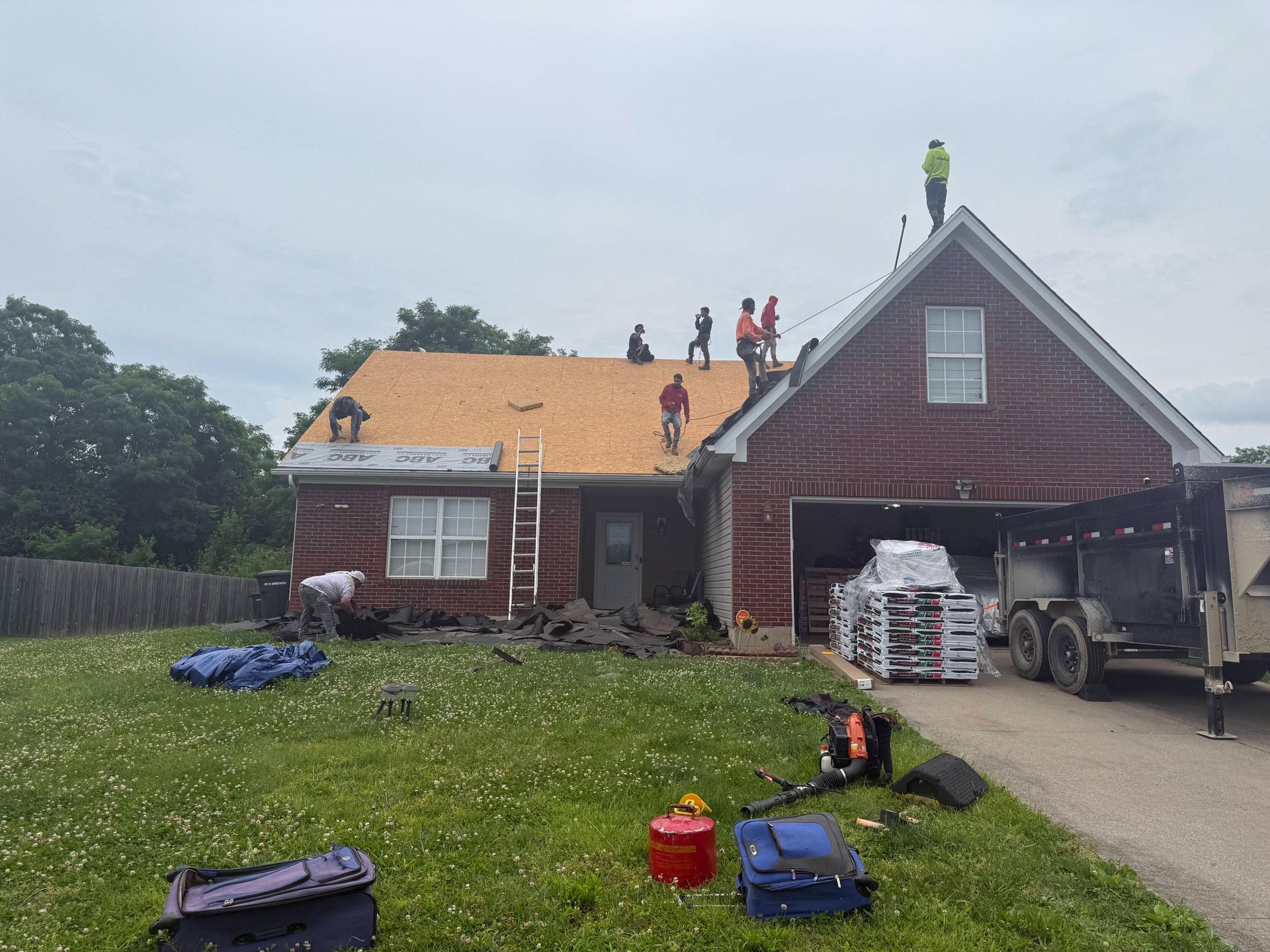 Workers repairing a red house roof, with shingles stacked in the driveway and yard tools on the lawn.