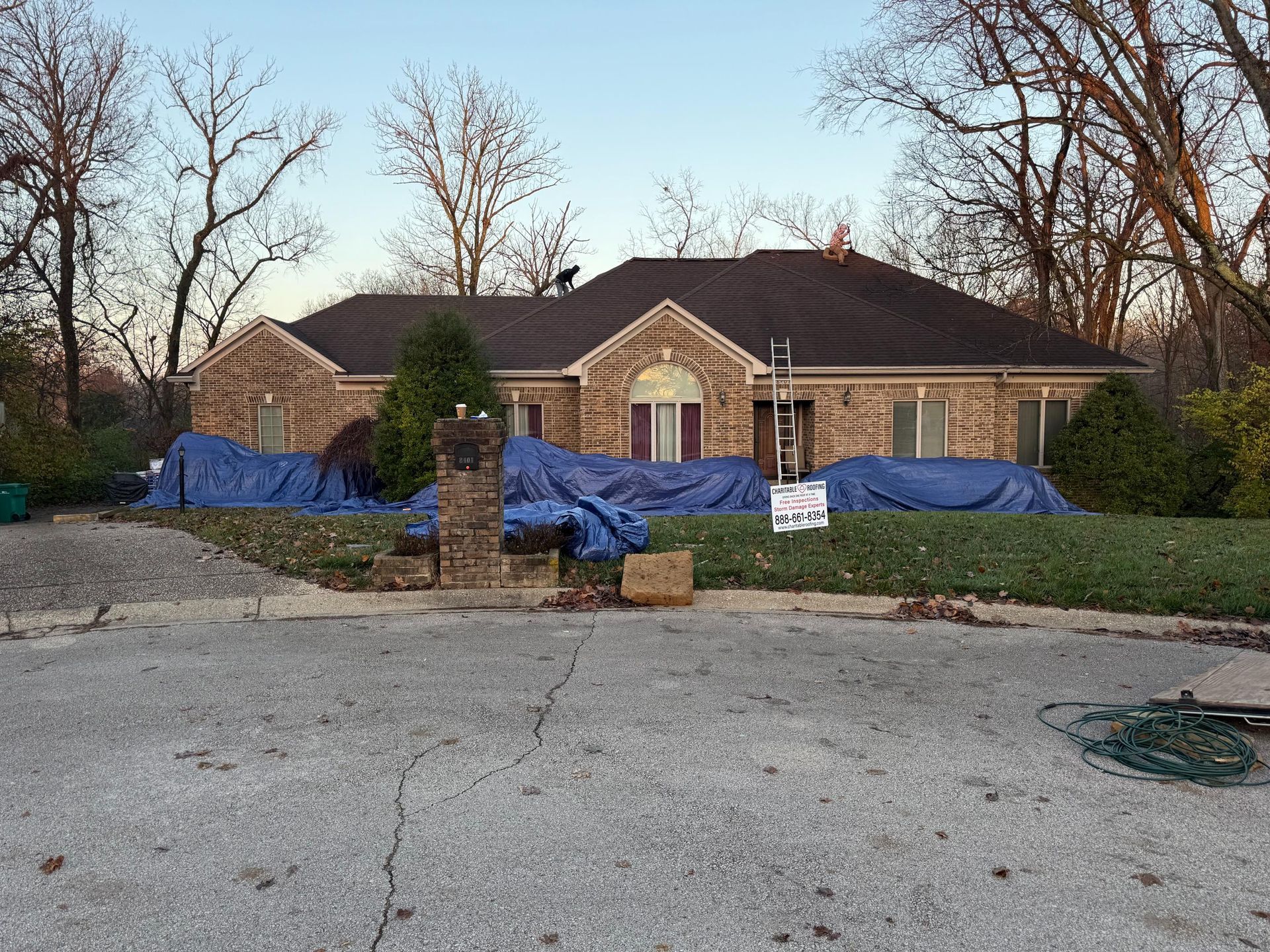 Brick house with roof damage covered by blue tarps behind a low stone wall and bare trees