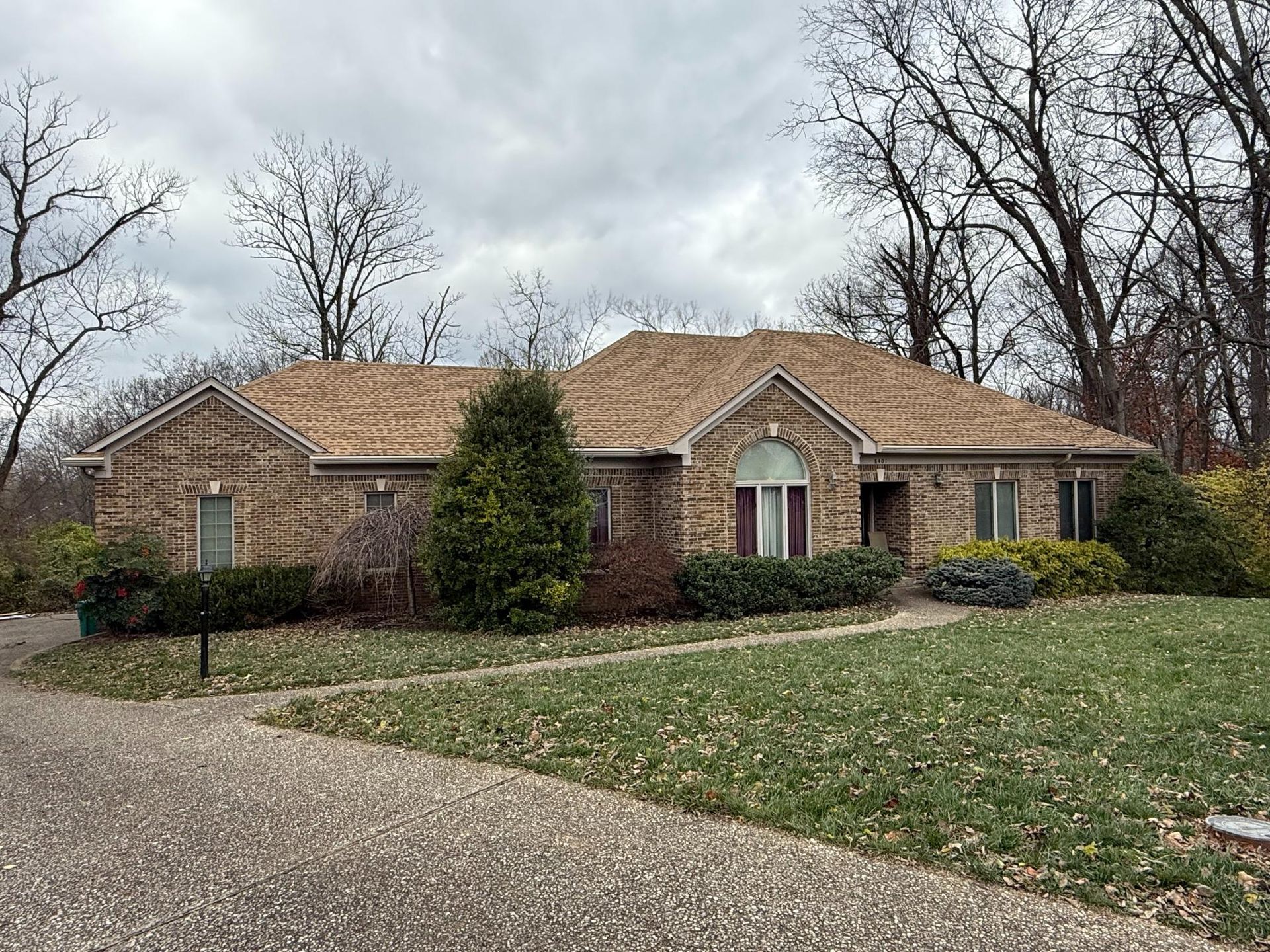 Brick suburban house with tan roof, front lawn, and leafless trees on a cloudy day