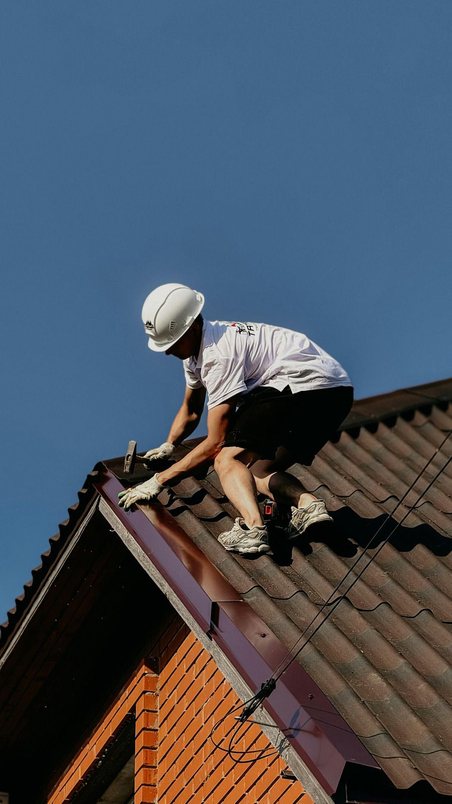 Worker on a roof repairing purple flashing beside brown tiles under a clear blue sky