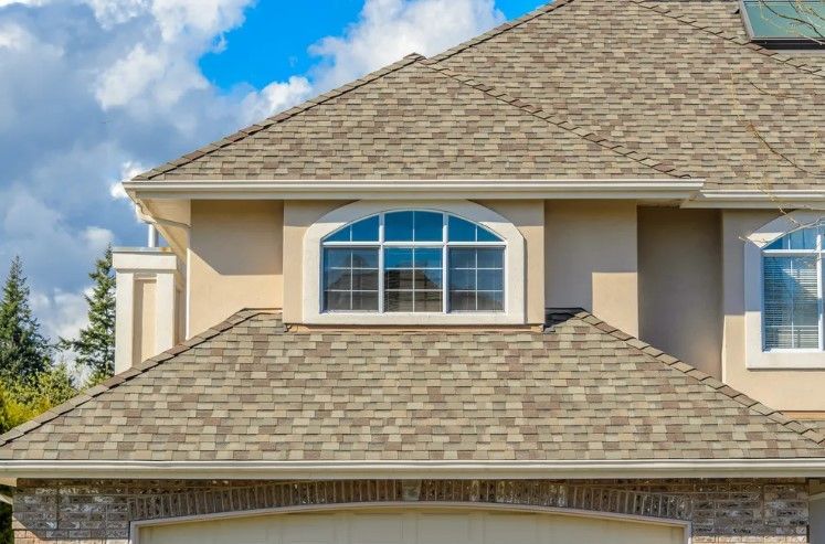 Suburban house roof and upper facade with beige shingles and arched window under blue sky