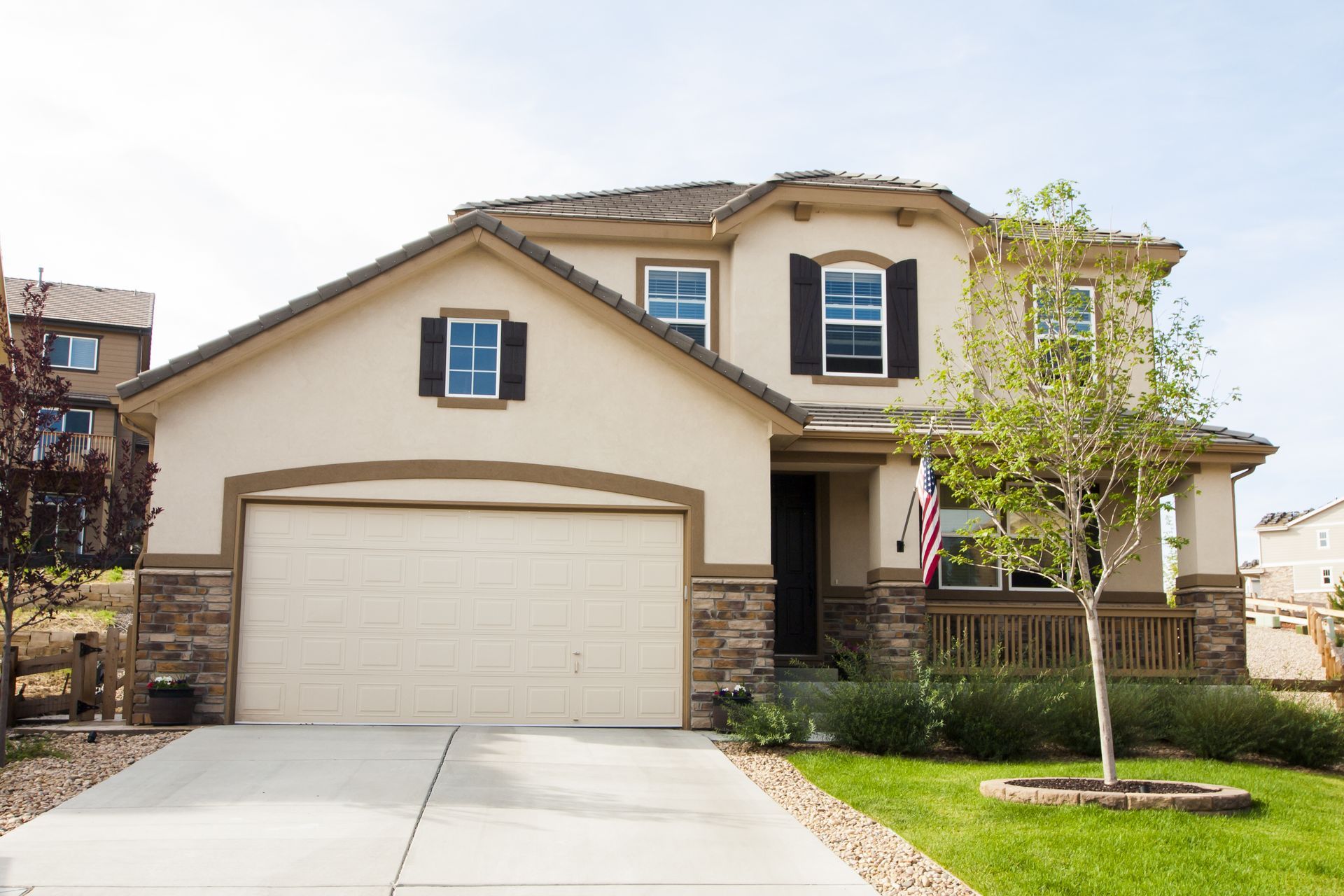 Suburban two-story beige house with a two-car garage, front porch, and a small tree by the lawn