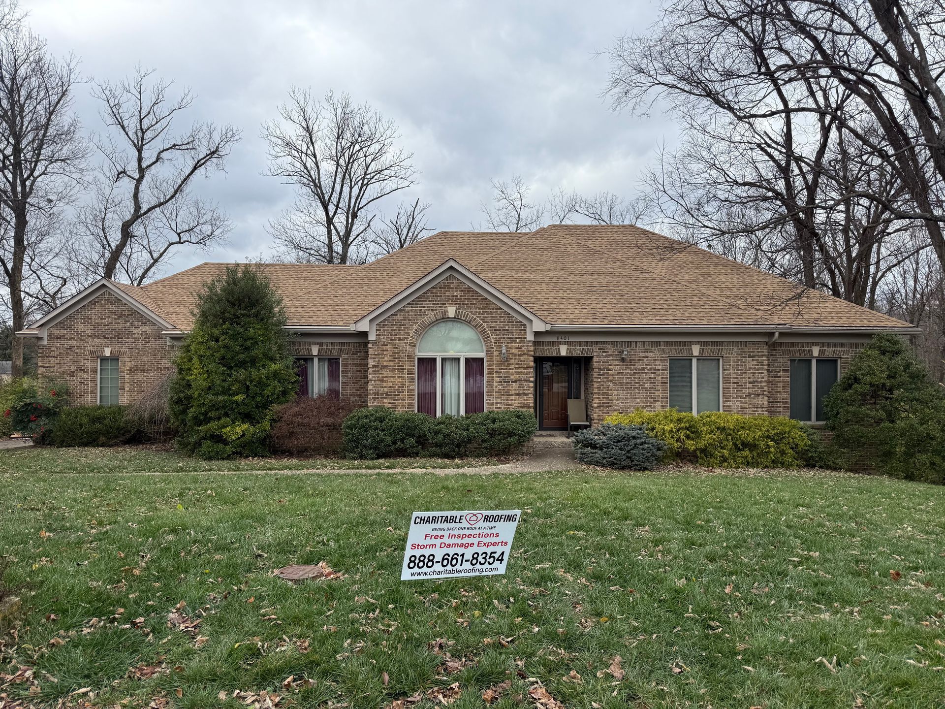 Single-story brick house with a tan roof, front lawn, and a small yard sign in front