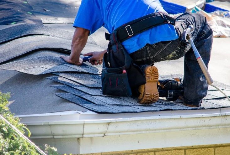 Worker in blue shirt installing shingles on a roof with a hammer and tool belt