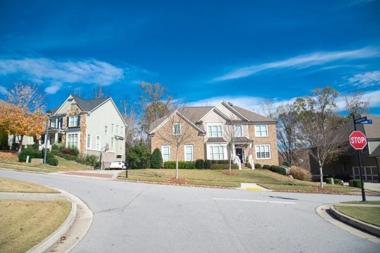 Suburban neighborhood street with beige houses, bare trees, and a stop sign under a blue sky