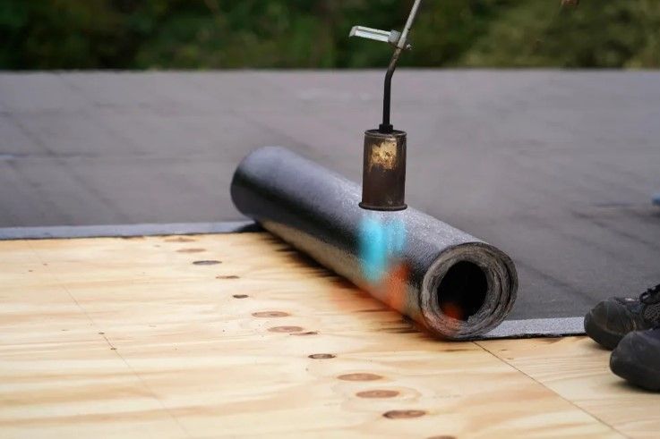 Torch heating a black roofing roll on a flat roof beside a wooden deck
