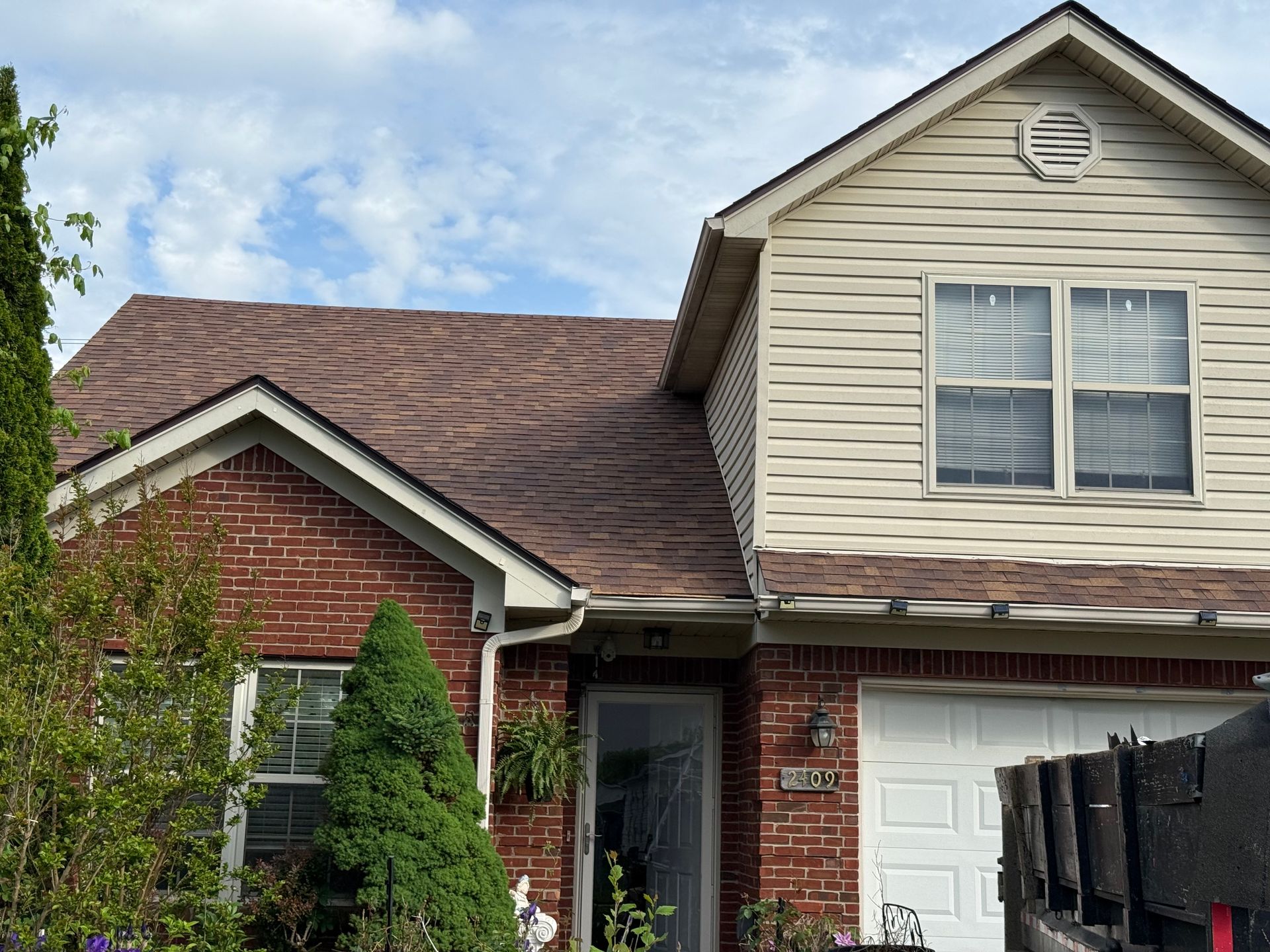 Suburban brick house with beige siding, white garage, and front porch under a partly cloudy sky