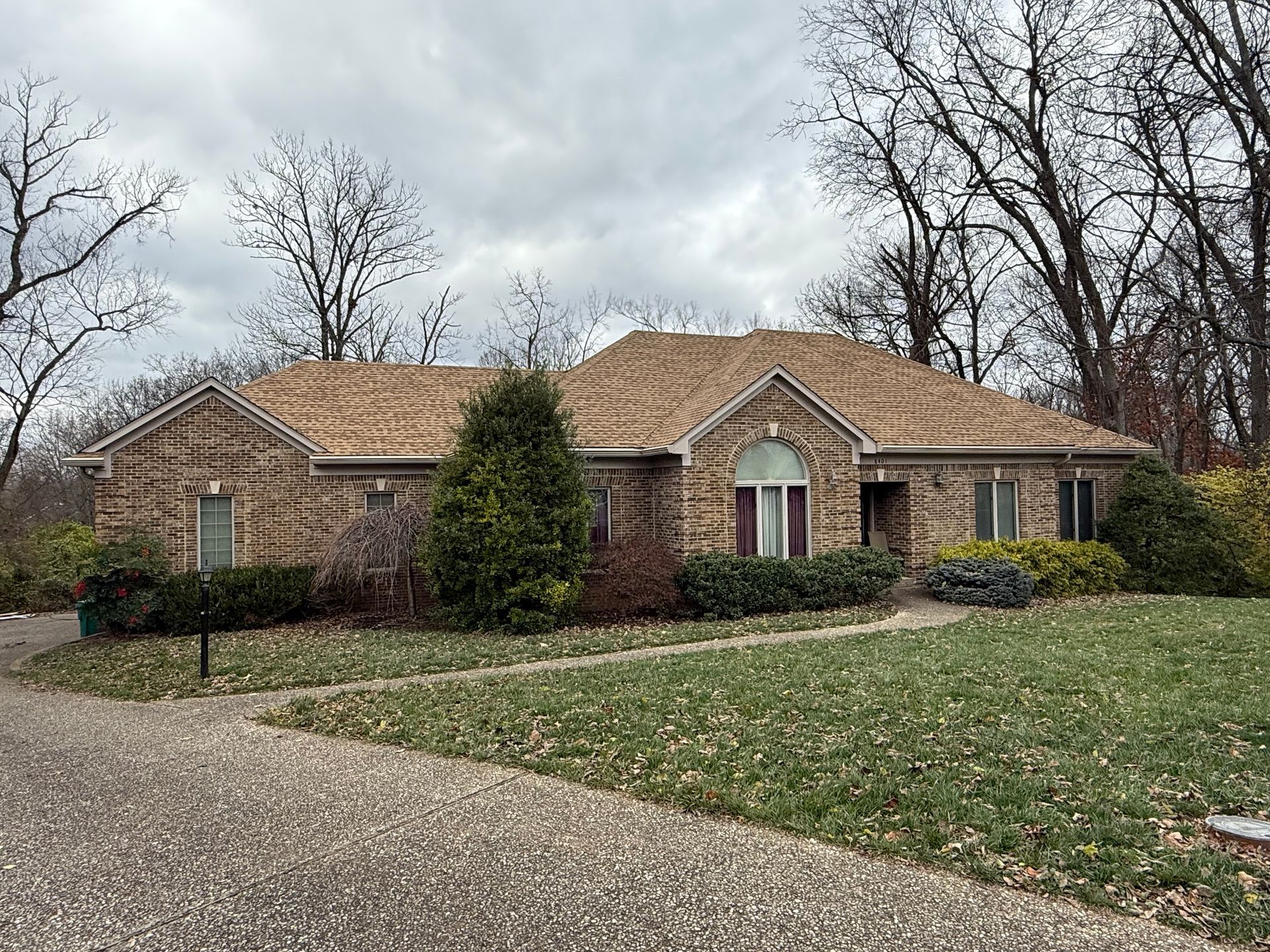 Suburban brick house with tan roof, driveway, and leaf-strewn lawn under a cloudy sky