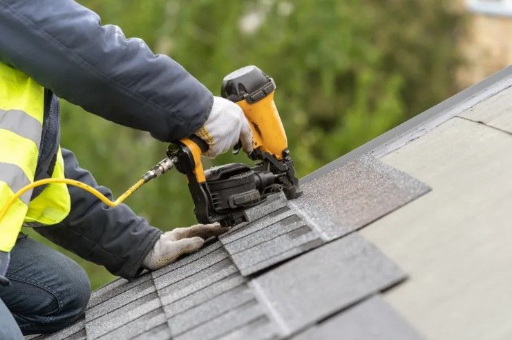 Worker using a yellow roofing nail gun to install gray shingles on a sloped roof