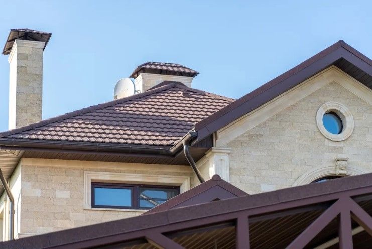 Stone house with brown tiled roof, chimney, and round window against a blue sky