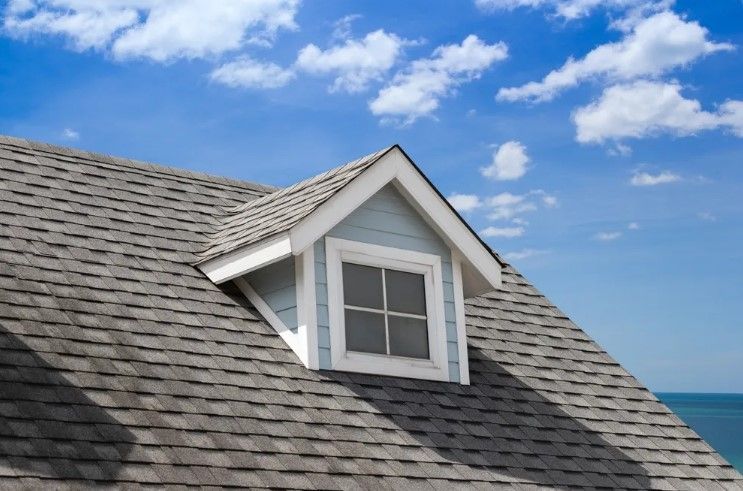 Dormer window on a gray shingled roof beside a blue ocean and cloudy sky