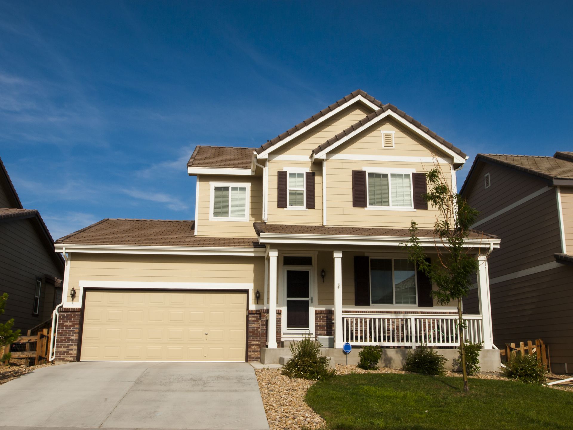Suburban two-story beige house with a garage, white porch, and blue sky.
