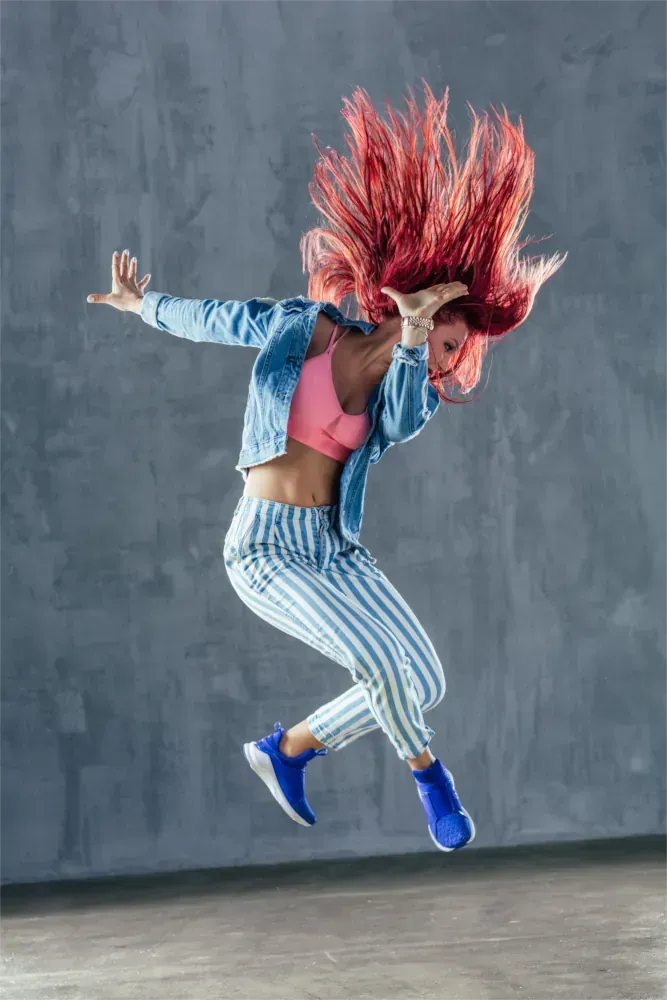 Eine Person springt gerade vor einer graue Wand; ihre roten Haare wehen im Wind; sie trägt eine blaue Jacke, eine gestreifte Hose und blaue Schuhe.