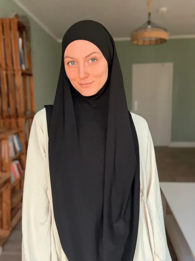 Eine Person in schwarzem Hijab und weißer Strickjacke steht in einem Raum mit grünen Wänden und lächelt sanft.