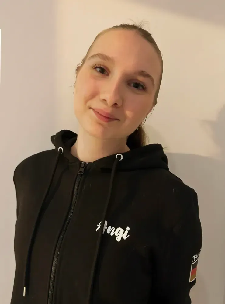 Eine Person in einem schwarzen Kapuzenpullover lächelt vor einer schlichten, hellen Wand.