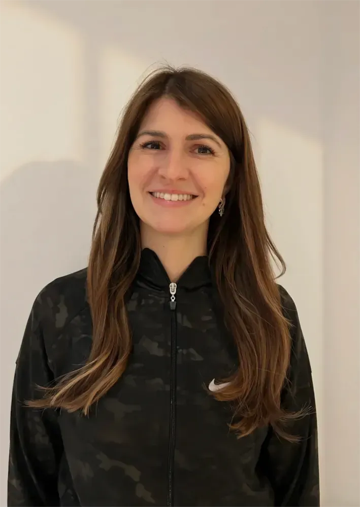 Eine lächelnde Frau in einer schwarzen Reißverschlussjacke steht vor einer hellen Wand.