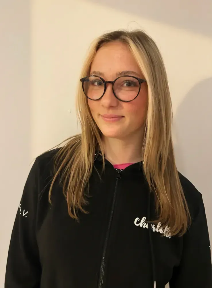 Eine Frau mit Brille und schwarzem Kapuzenpullover steht vor einer schlichten beigen Wand.
