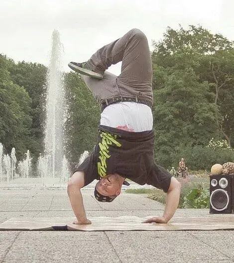 Eine Person macht einen Handstand im Bereich eines Parkbrunnens; im Hintergrund sind Bäume und ein Lautsprecher zu sehen.