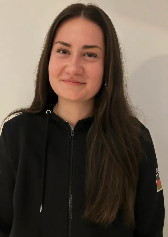 Person mit langen dunklen Haaren, die einen schwarzen Kapuzenpullover trägt, steht vor einer hellen Wand und lächelt sanft.