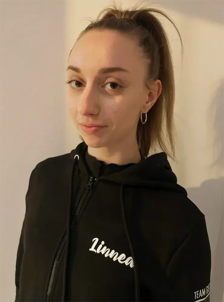 Person in schwarzem Kapuzenpulli mit Pferdeschwanz, die drinnen vor einer hellen Wand steht.