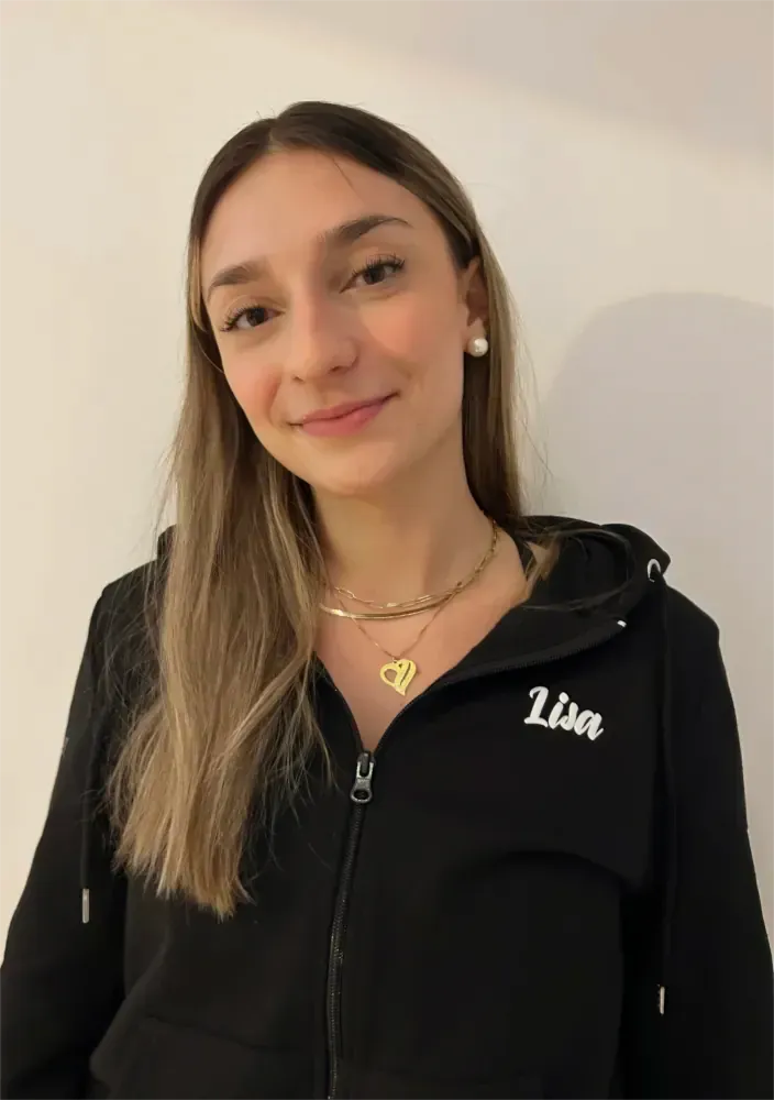 Person lächelt in schwarzem Kapuzenpulli und mit Halskette vor einer schlichten Wand.