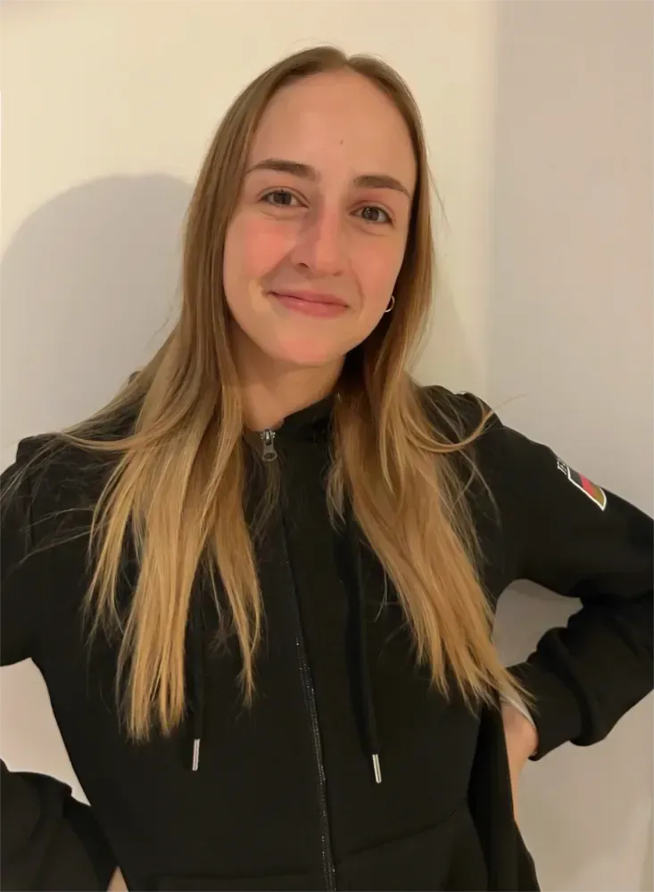 Person in einer schwarzen Reißverschlussjacke lächelt vor einer schlichten, hellen Wand.