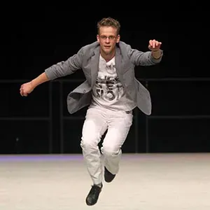 Eine Person in einem grauen Blazer und einer weißen Hose springt auf einer dunklen Bühne auf die Kamera zu.