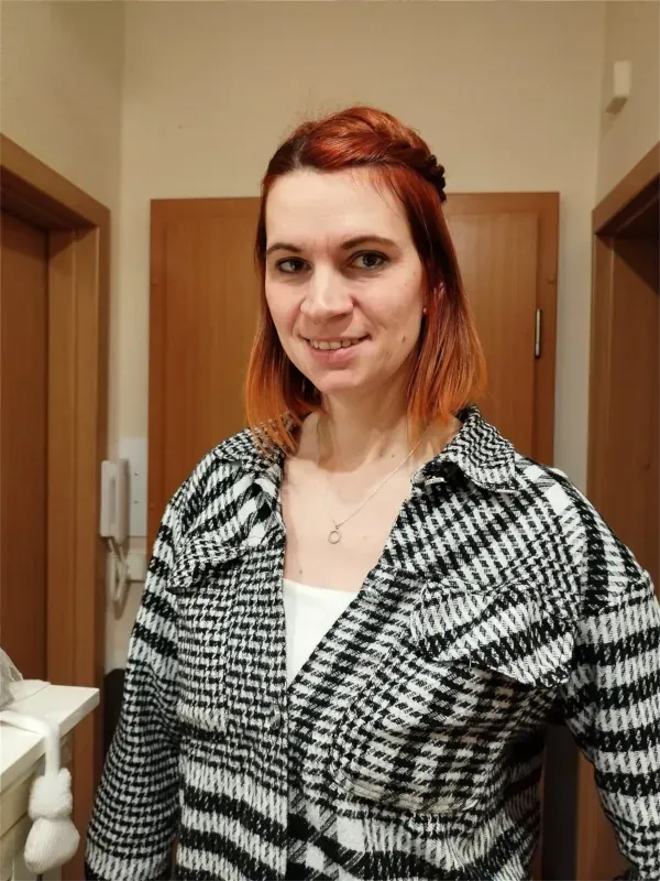 Eine rothaarige Frau lächelt in einem Flur; sie trägt ein schwarz-weiß gemustertes Hemd.