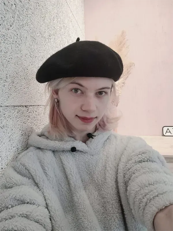 Eine Person mit schwarzer Baskenmütze und grauem, flauschigem Pullover macht ein Selfie vor einer hellen Wand.