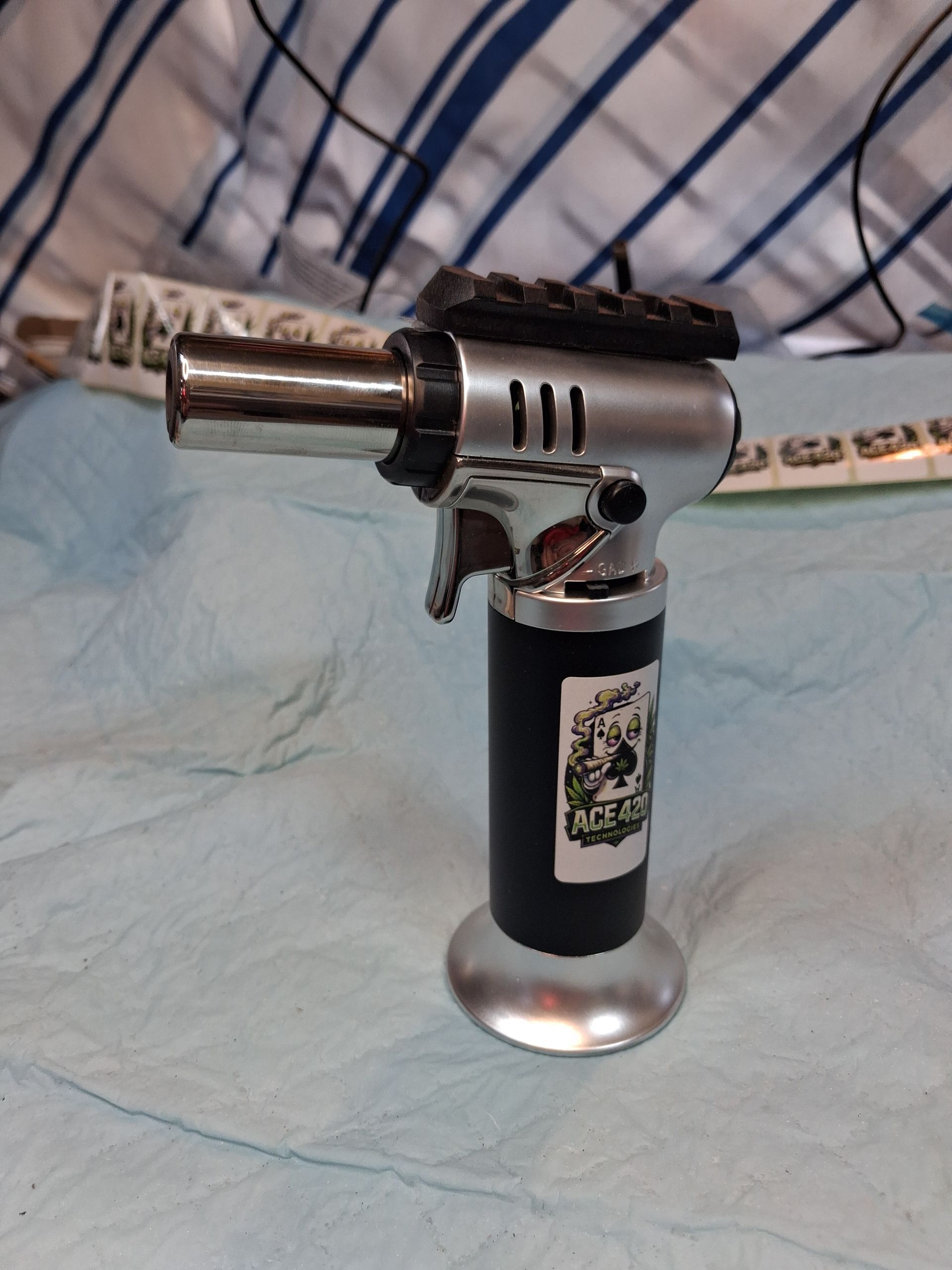 Metal torch lighter standing on a blue cloth with a row of stickers in the background.