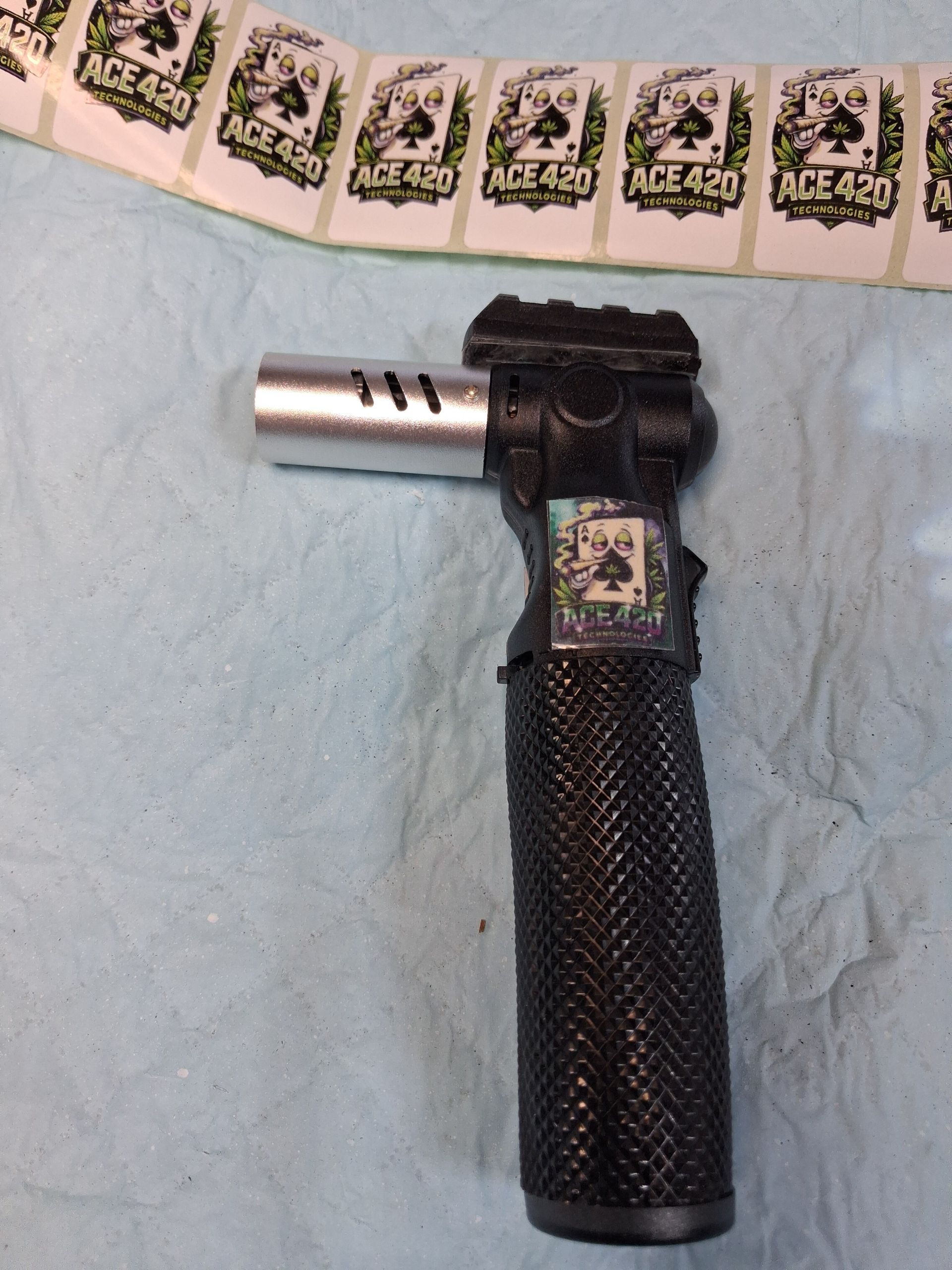 Black handheld blowtorch-like tool with a long textured grip on a light blue surface, near wrapped packets.