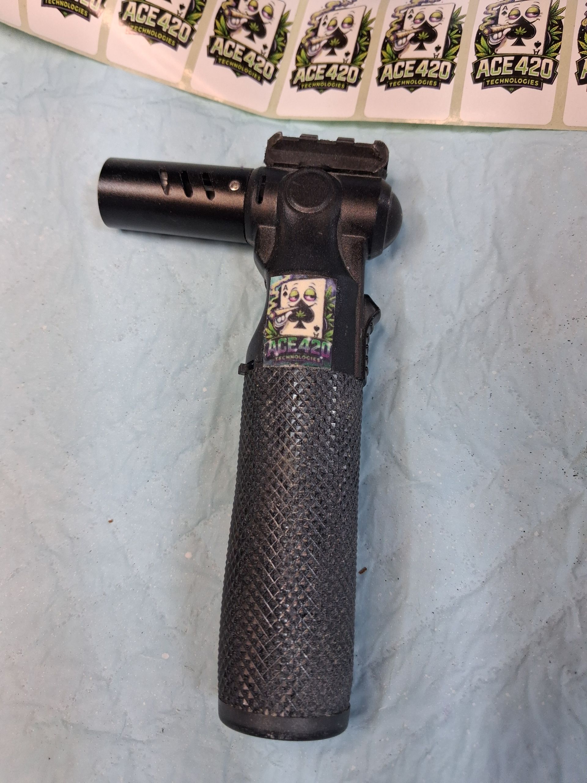 Black handheld blowtorch-like tool with a long textured grip on a light blue surface, near wrapped packets.