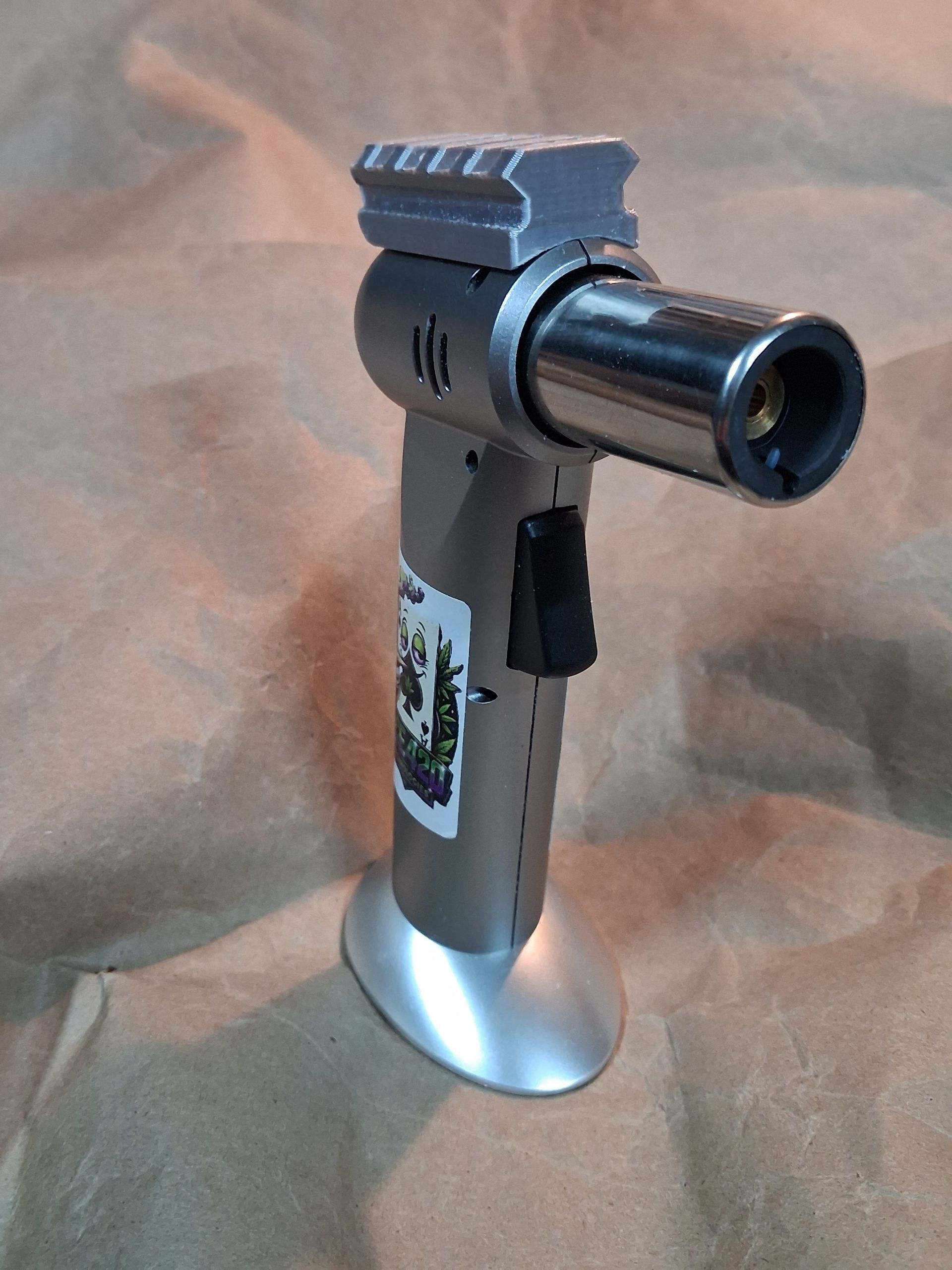 Silver butane torch lighter with black trigger and decorative label on a tan surface