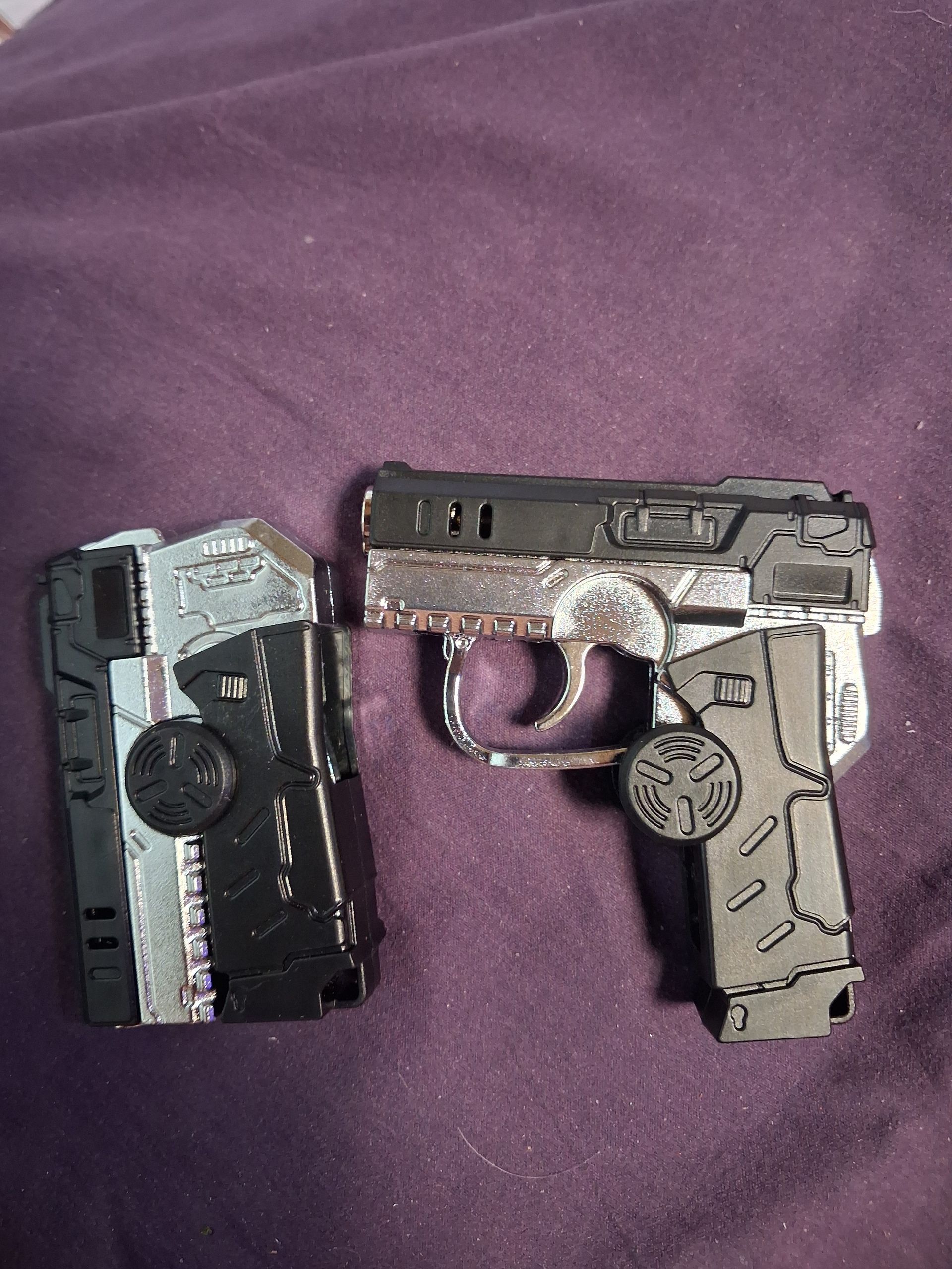 Two toy pistols, one black and silver, one tan and silver, on a purple fabric background