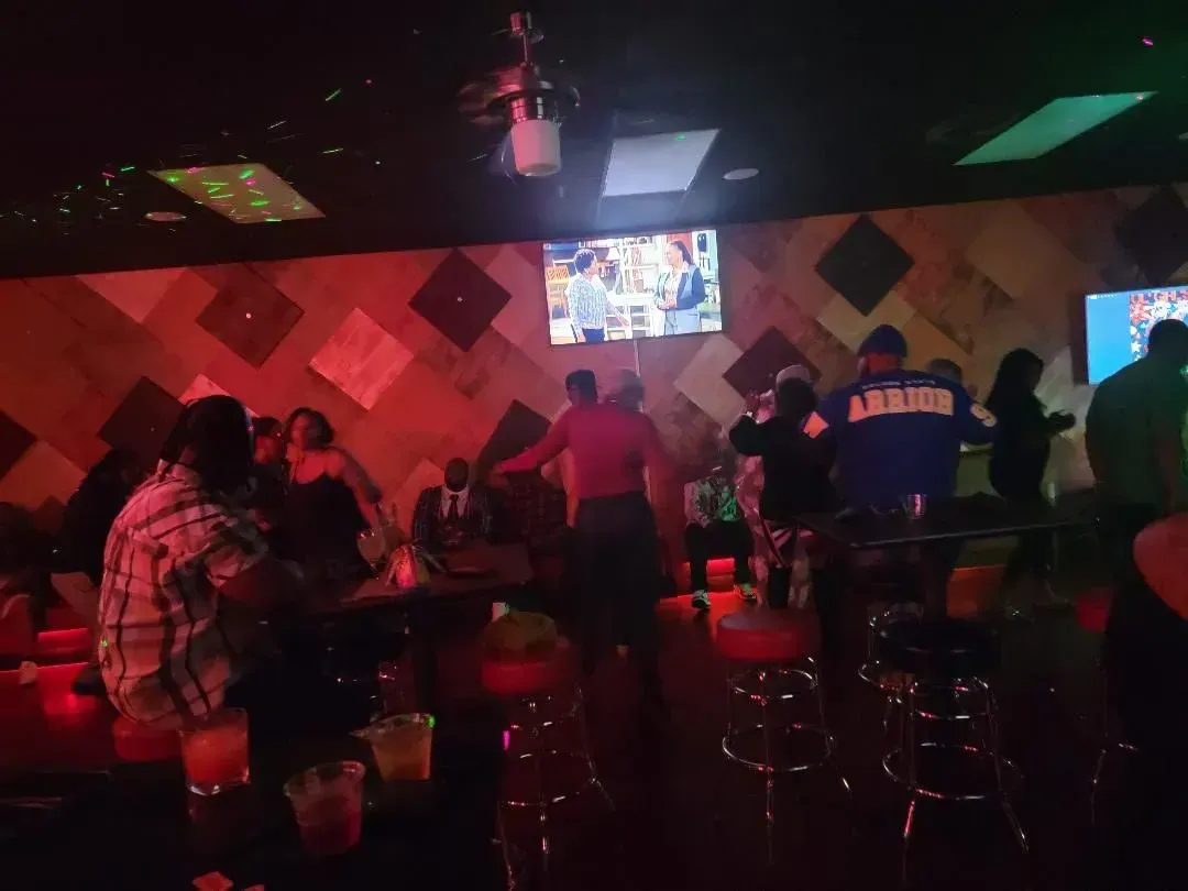 Dimly lit bar with patrons seated at tables, TVs on the wall, and red ambient lighting