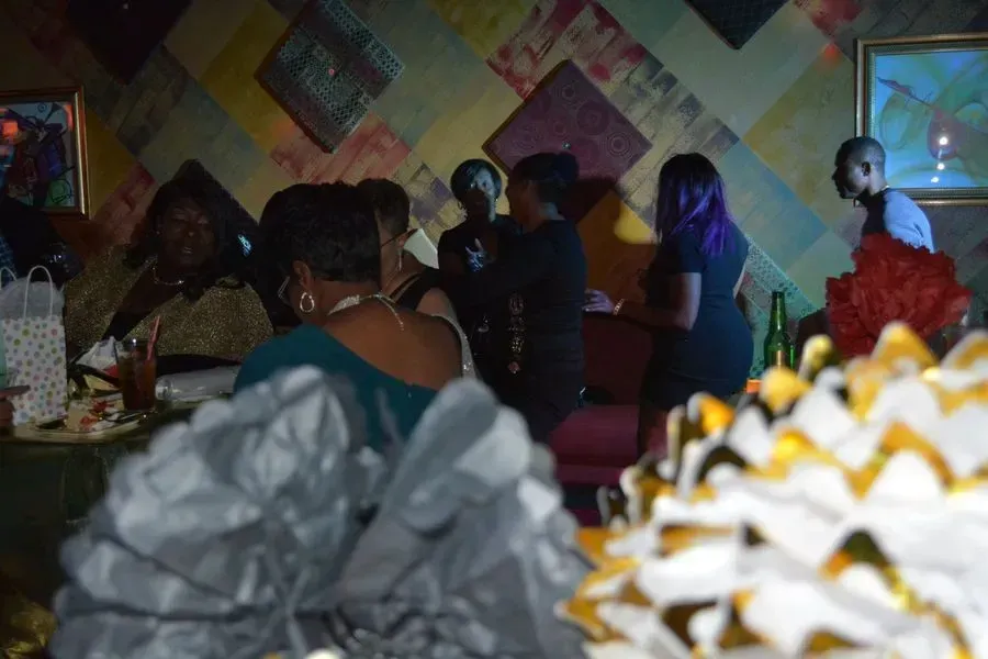 Dim indoor party scene with people mingling near a decorated wall and white-and-yellow floral arrangements in front.